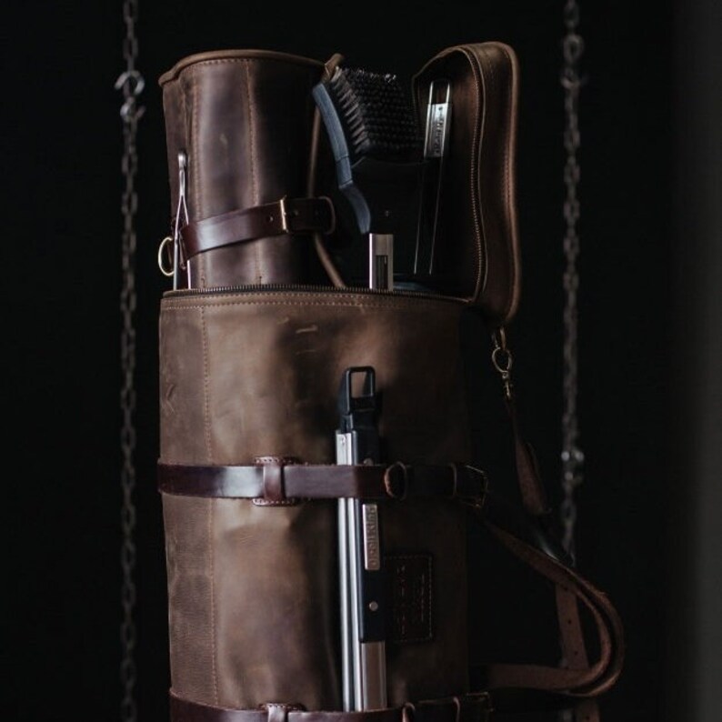 May include: A brown leather bag with a zippered top and multiple straps. The bag is filled with various tools, including a brush, a metal tool, and a long, thin metal tool.