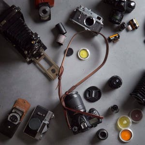 May include: A collection of vintage and modern cameras, lenses, and film canisters on a gray surface. The cameras include a large format camera, a rangefinder camera, and a digital SLR camera. The lenses are a variety of sizes and shapes, and the film canisters are both full and empty.