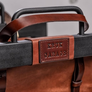 May include: A brown leather bag with a black metal frame and a brown leather handle. The bag has a leather patch with the text "KRUK GARAGE" embossed on it.