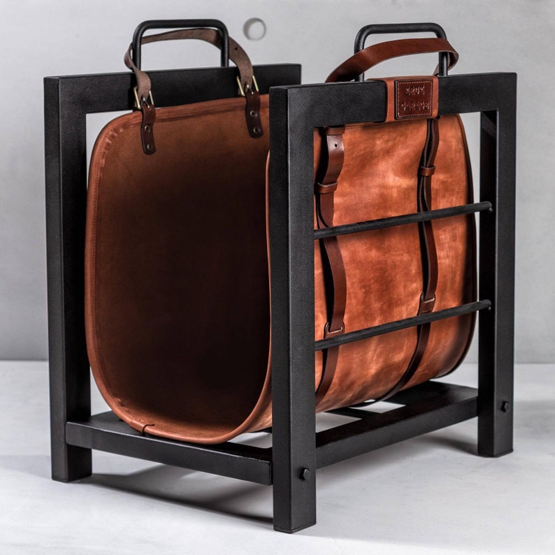 May include: A black metal log holder with a brown leather log basket. The basket is made of two pieces of leather that are connected by metal straps. The log holder has a rectangular shape and is standing on a white surface.