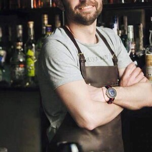 May include: A man wearing a gray t-shirt with a brown leather apron and a watch on his wrist. The apron has a strap that goes over the shoulder and a buckle.