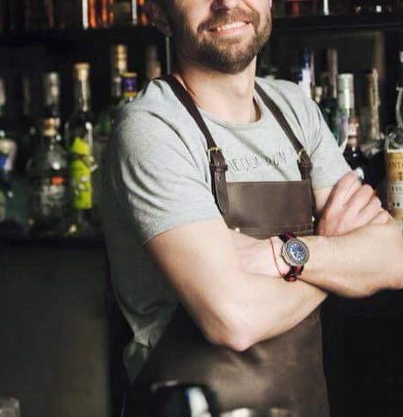 May include: A man wearing a gray t-shirt with a brown leather apron and a watch on his wrist. The apron has a strap that goes over the shoulder and a buckle.