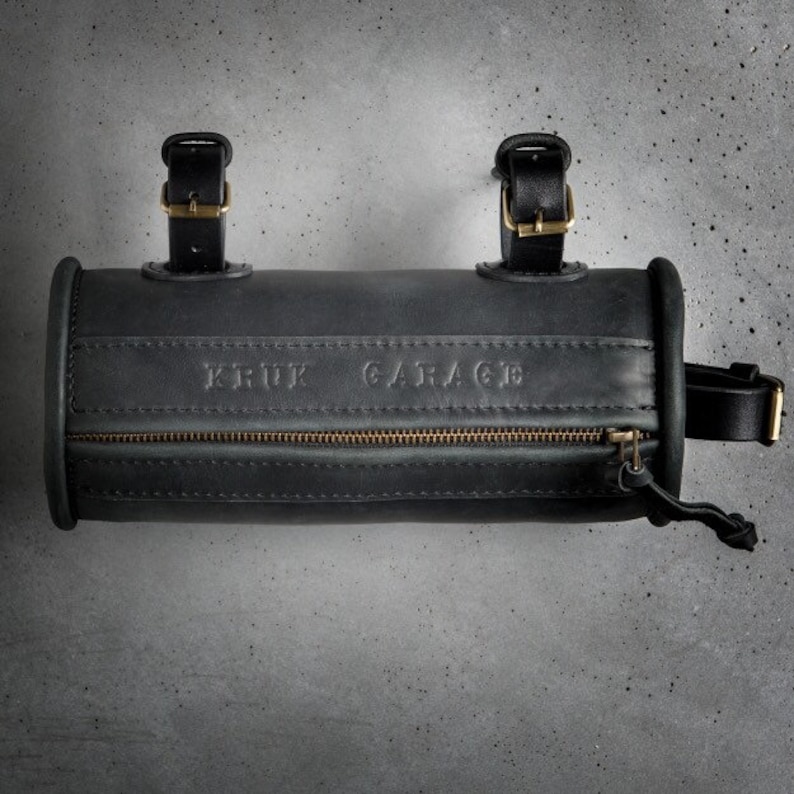May include: A black leather handlebar bag with a zipper closure and a brass buckle. The bag has the text "KRUK GARAGE" embossed on the front.