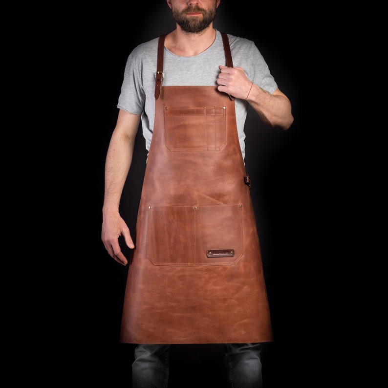 May include: A brown leather apron with adjustable straps and multiple pockets. The apron is worn by a person with a beard and a grey shirt.