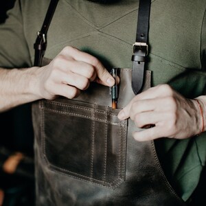 May include: A brown leather apron with a large pocket and a pen tucked into the pocket. The apron has adjustable straps.