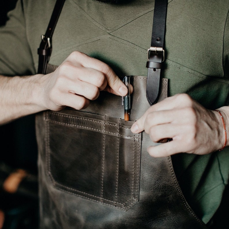 May include: A brown leather apron with a large pocket and a pen tucked into the pocket. The apron has adjustable straps.