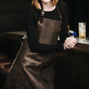 May include: A brown leather apron with adjustable straps and a large pocket. The apron has the text "Anus Garage" embossed on the front.