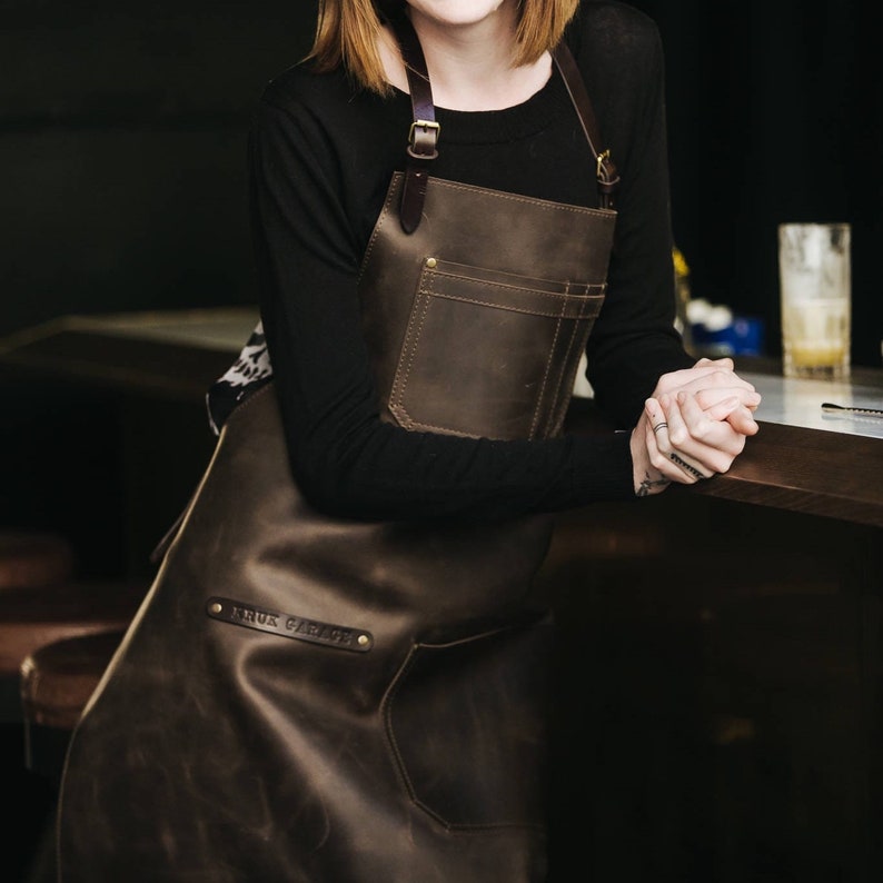 May include: A brown leather apron with adjustable straps and a large pocket. The apron has the text "Anus Garage" embossed on the front.