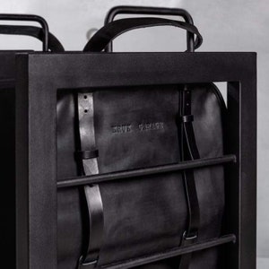 May include: A black leather backpack with adjustable straps hanging in a black metal frame. The backpack has the text "BRUK GARAGE" embossed on the front.