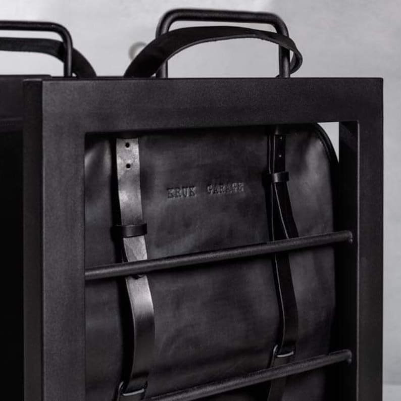 May include: A black leather backpack with adjustable straps hanging in a black metal frame. The backpack has the text "BRUK GARAGE" embossed on the front.