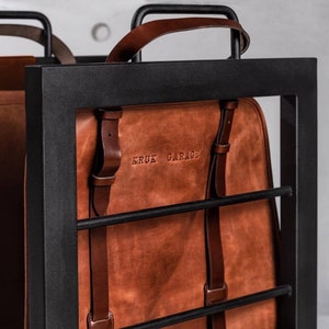 May include: A brown leather backpack with black straps and the text "KRUK GARAGE" embossed on the front. The backpack is hanging on a black metal rack.