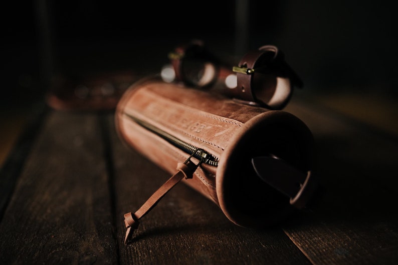 May include: A brown leather cylindrical pouch with a zipper closure and a leather strap. The pouch is lying on a wooden surface.