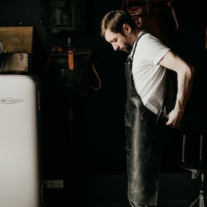May include: A man wearing a black leather apron in a workshop setting. The apron has metal hardware and is worn over a white t-shirt. A vintage white refrigerator and leather bags are in the background.