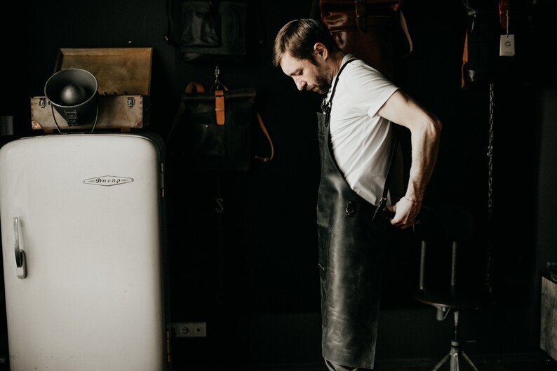 May include: A man wearing a black leather apron in a workshop setting. The apron has metal hardware and is worn over a white t-shirt. A vintage white refrigerator and leather bags are in the background.