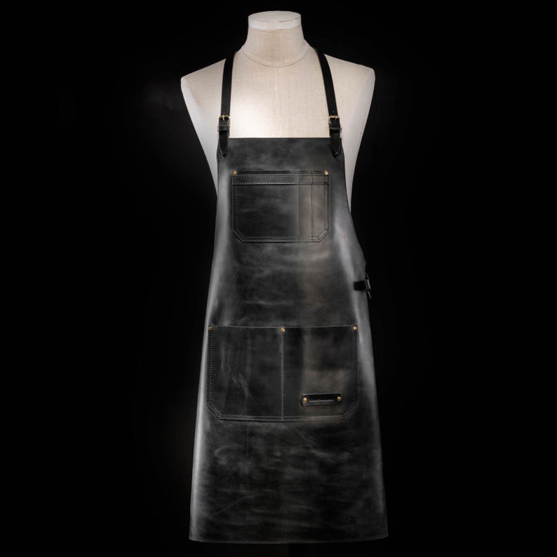 May include: A dark gray leather apron with two large pockets and brass rivets. The apron has adjustable straps and a buckle closure. The apron is displayed on a mannequin against a black background.