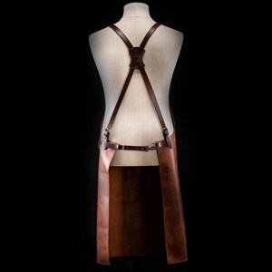 May include: Brown leather apron with adjustable straps and a buckle closure. The apron has a long, rectangular shape and is designed for use in a kitchen or workshop.
