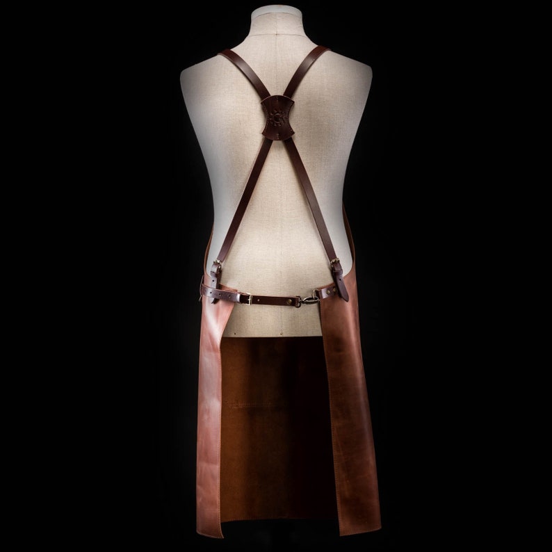 May include: Brown leather apron with adjustable straps and a buckle closure. The apron has a long, rectangular shape and is designed for use in a kitchen or workshop.