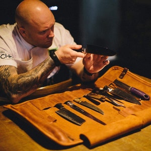 May include: A brown leather knife roll with multiple slots for knives. The roll is unrolled and several knives are visible. A person is holding a black knife in their hands.