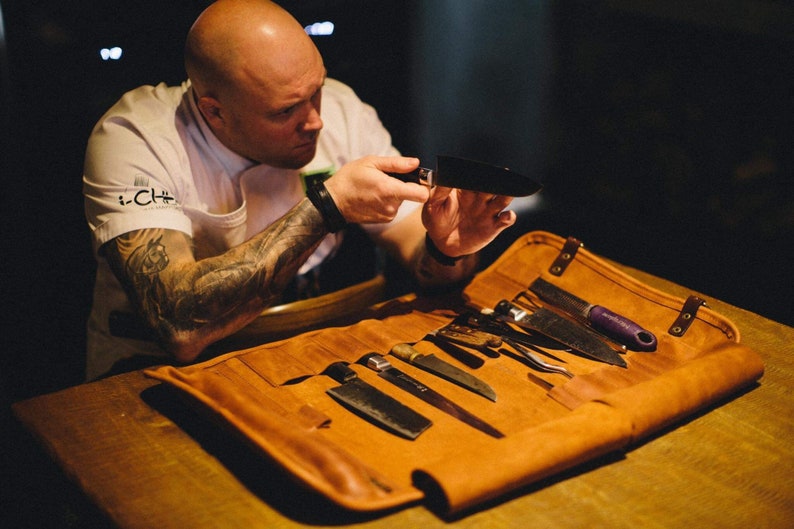 May include: A brown leather knife roll with multiple slots for knives. The roll is unrolled and several knives are visible. A person is holding a black knife in their hands.