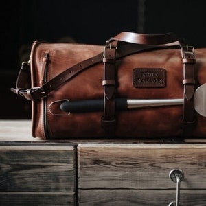 May include: A brown leather duffel bag with a long, silver metal tool strapped to the side. The bag has a leather strap with the words "ROTHCO" and "GARAGE" embossed on it.