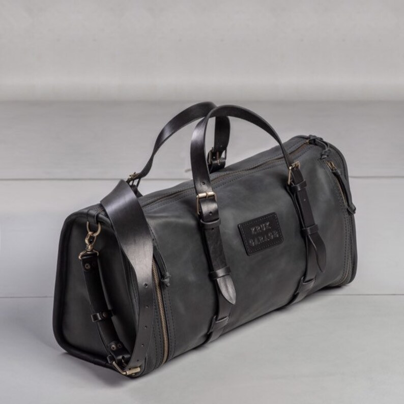 May include: Black leather duffel bag with multiple straps and buckles. The bag has a zipper closure and a label that reads "KRUX GARAGE".