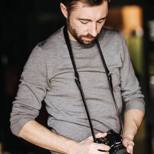 May include: A man wearing a gray sweater and jeans is holding a black camera with a black leather strap. The camera has a lens with a silver ring around it.