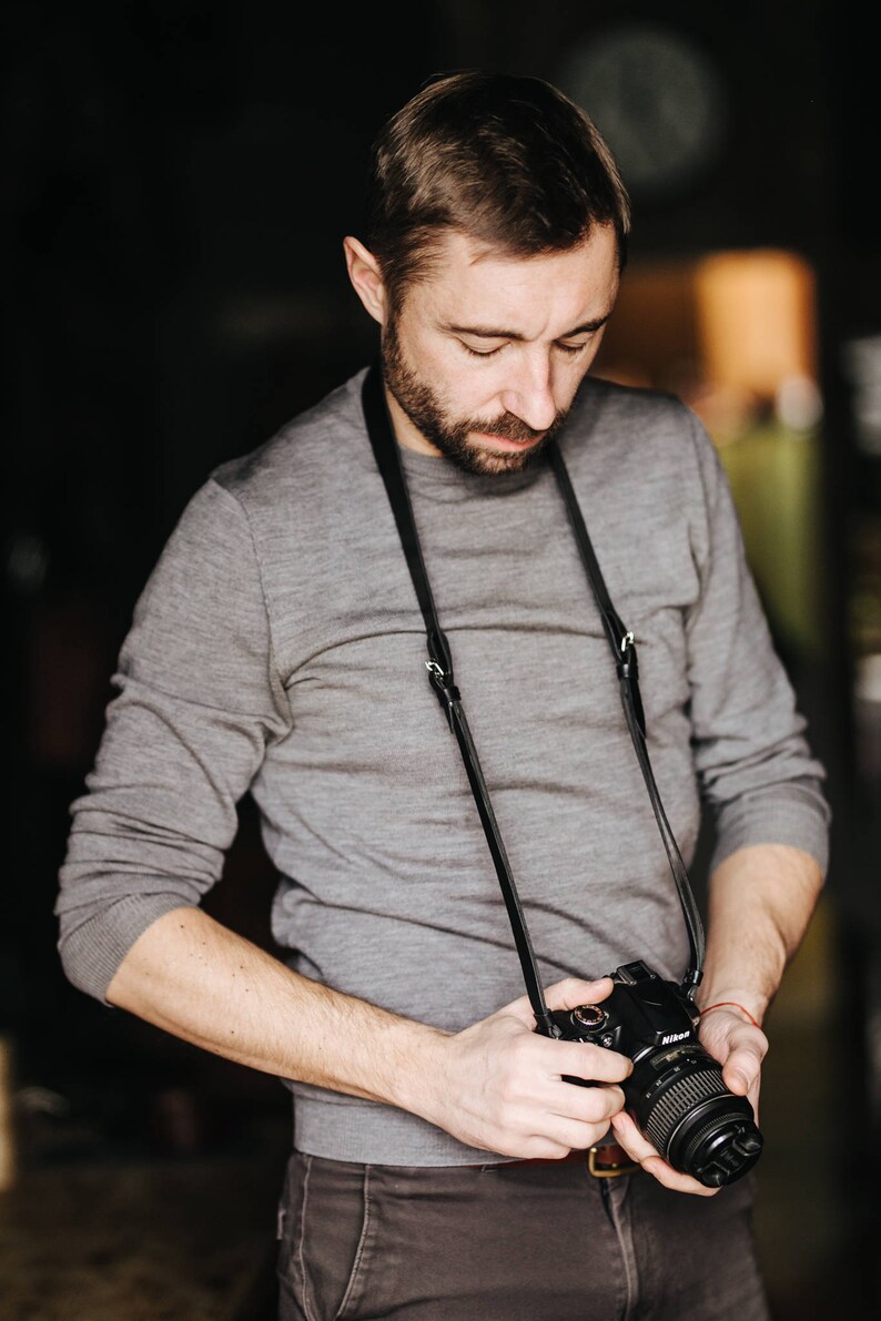 May include: A man wearing a gray sweater and jeans is holding a black camera with a black leather strap. The camera has a lens with a silver ring around it.