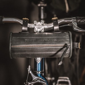 May include: A black leather handlebar bag with a zipper and the text "KRUK GARAGE" embossed on the front. The bag is attached to a bicycle handlebar.