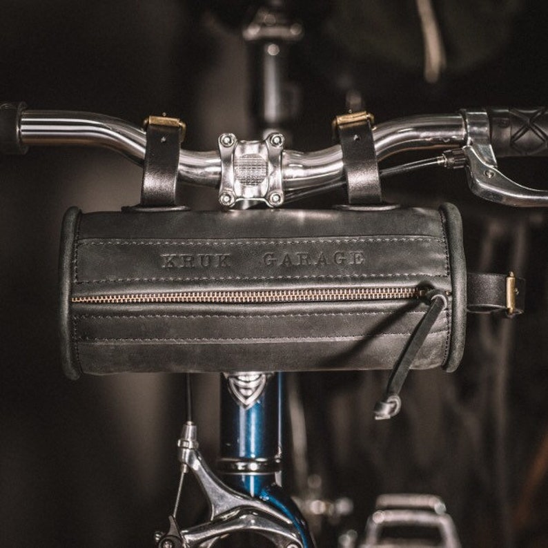 May include: A black leather handlebar bag with a zipper and the text "KRUK GARAGE" embossed on the front. The bag is attached to a bicycle handlebar.