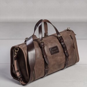 May include: A brown leather duffel bag with a dark brown leather strap and buckles. The bag has a zipper closure and a small leather patch with the text "RINK" embossed on it.