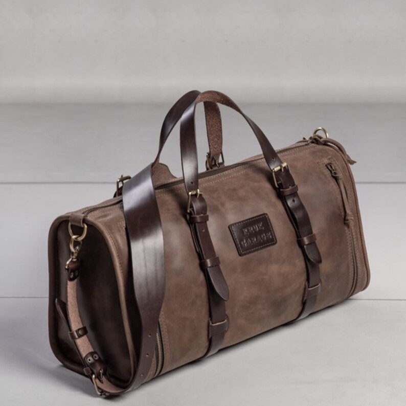 May include: A brown leather duffel bag with a dark brown leather strap and buckles. The bag has a zipper closure and a small leather patch with the text "RINK" embossed on it.