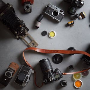 May include: A flat lay of various vintage and modern cameras, film canisters, and camera accessories on a gray concrete surface. The cameras include a black and silver rangefinder camera, a black and silver SLR camera, a black and silver view camera, and a black and silver digital SLR camera. The accessories include a brown leather camera strap, a black lens cap, and a black film canister.