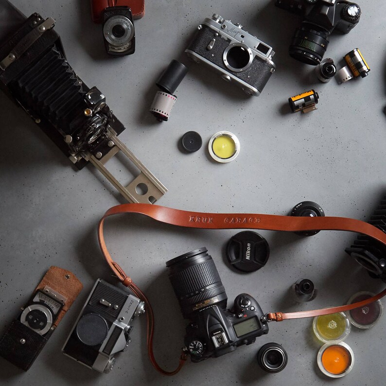 May include: A flat lay of various vintage and modern cameras, film canisters, and camera accessories on a gray concrete surface. The cameras include a black and silver rangefinder camera, a black and silver SLR camera, a black and silver view camera, and a black and silver digital SLR camera. The accessories include a brown leather camera strap, a black lens cap, and a black film canister.