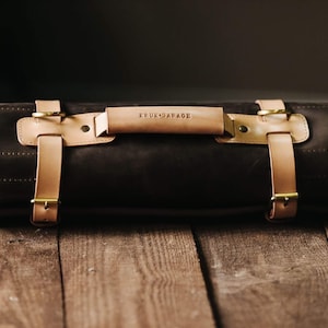 May include: A brown leather roll-up bag with tan leather straps and brass buckles. The bag has a tan leather handle with the text "KRUX GARAGE" embossed on it.