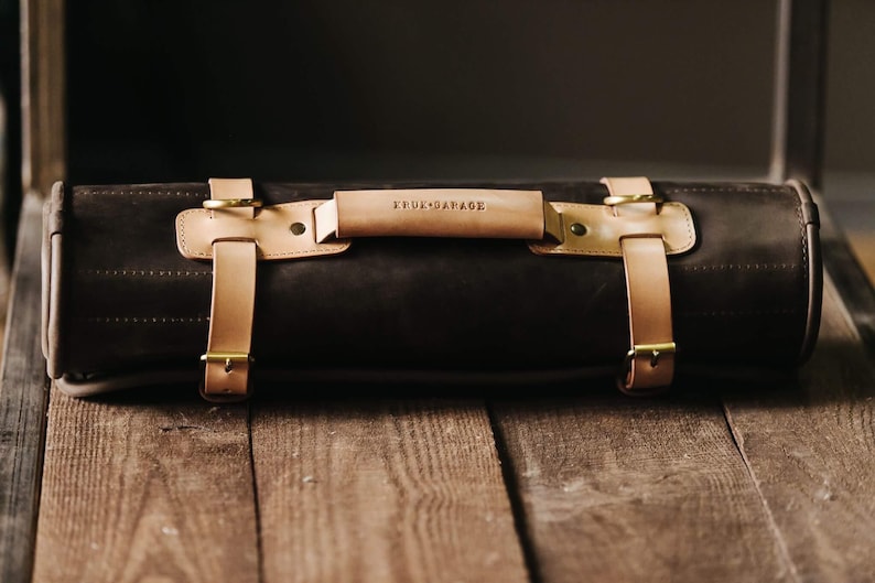 May include: A brown leather roll-up bag with tan leather straps and brass buckles. The bag has a tan leather handle with the text "KRUX GARAGE" embossed on it.