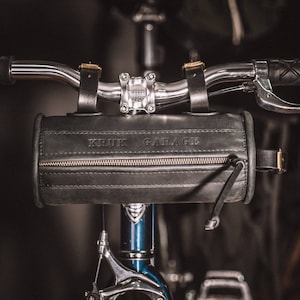 May include: A black leather handlebar bag with a zipper and the text "KRUK GARAGE" embossed on the front. The bag is attached to a bicycle handlebar.