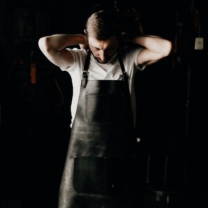 May include: A black leather apron with two large pockets and adjustable straps. The apron is worn over a white t-shirt. The image is set against a dark background, highlighting the apron's design and texture.