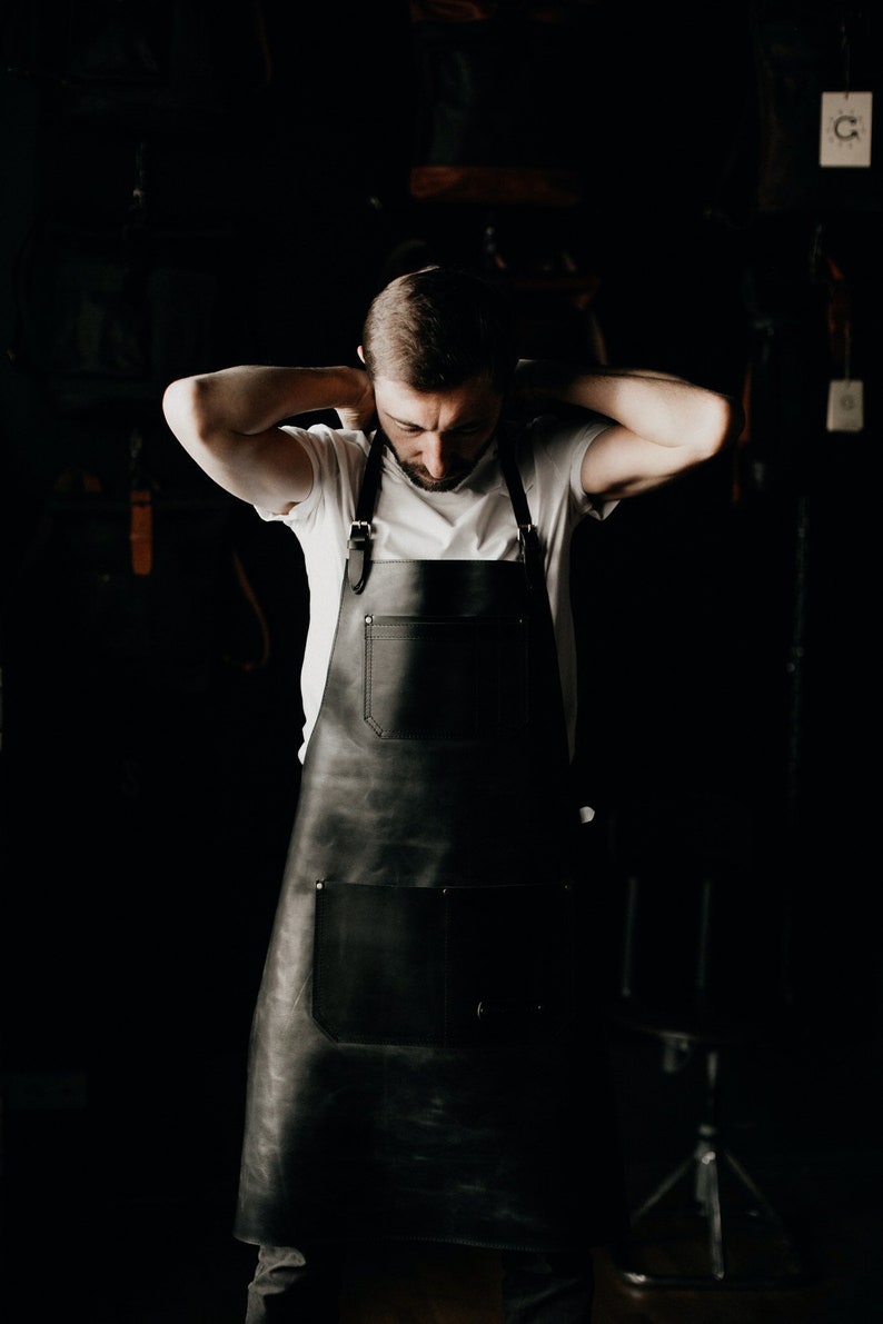May include: A black leather apron with two large pockets and adjustable straps. The apron is worn over a white t-shirt. The image is set against a dark background, highlighting the apron's design and texture.