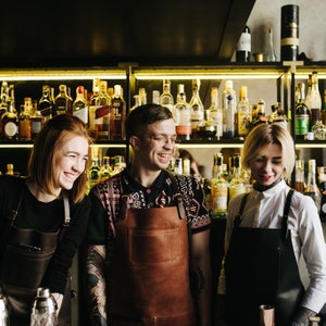 May include: Three people wearing aprons stand behind a bar with shelves of liquor bottles. The person in the middle is wearing a brown leather apron and a patterned shirt. The person on the left is wearing a black apron and a black shirt. The person on the right is wearing a black apron and a white shirt.