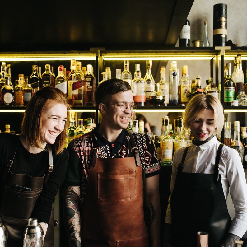 May include: Three people wearing aprons stand behind a bar with shelves of liquor bottles. The person in the middle is wearing a brown leather apron and a patterned shirt. The person on the left is wearing a black apron and a black shirt. The person on the right is wearing a black apron and a white shirt.