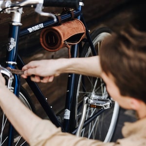 May include: A person adjusts a brown leather handlebar bag on a blue bicycle with silver handlebars and a silver seat. The bicycle has the text "PURE FIX" on the frame.