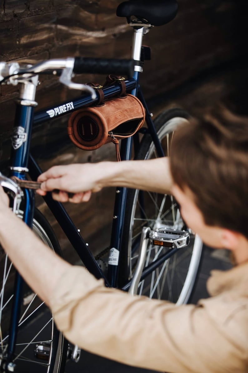 May include: A person adjusts a brown leather handlebar bag on a blue bicycle with silver handlebars and a silver seat. The bicycle has the text "PURE FIX" on the frame.