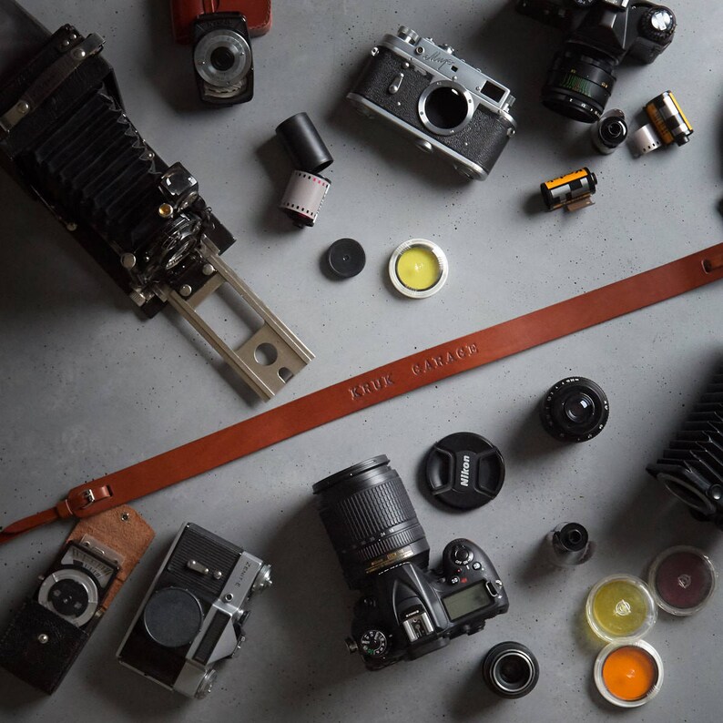 May include: A collection of vintage and modern cameras, lenses, and film rolls on a gray surface. A brown leather camera strap with the text "KRUK GARAGE" is visible in the image.