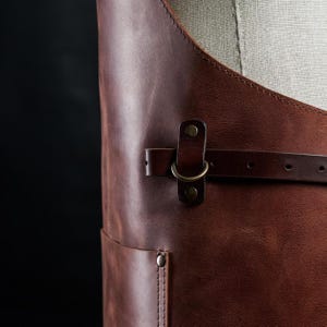 May include: A brown leather apron with a metal buckle and a strap. The apron has a large pocket on the front.
