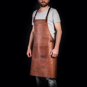 May include: A brown leather apron with adjustable straps and multiple pockets. The apron is worn by a person with a beard and a grey t-shirt.