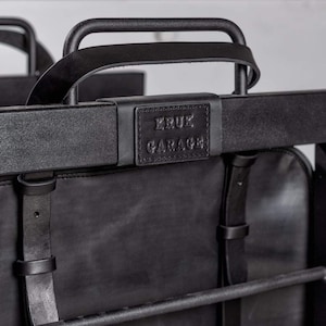 May include: Black leather backpack with a metal frame and a leather handle. The backpack has a black leather patch with the text "KRUK GARAGE" on it.