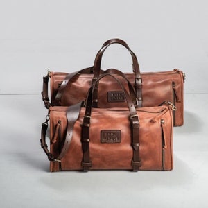 May include: Two brown leather travel bags with the text "True Garage" embossed on the front. The larger bag is behind the smaller bag.