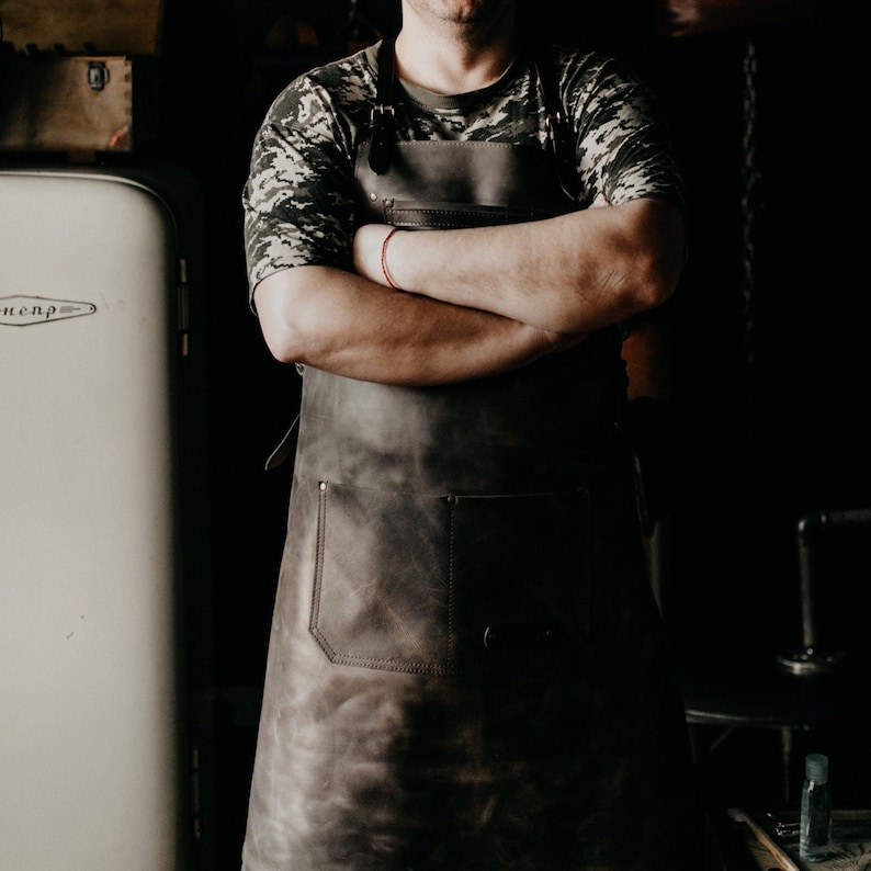 May include: A person wearing a camouflage shirt and a brown leather apron with a large pocket. The apron has a strap that goes around the neck and ties at the waist.