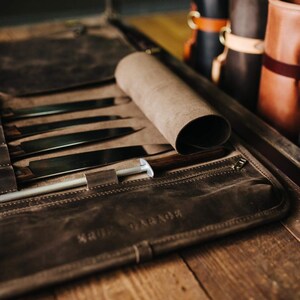 May include: A brown leather knife roll with a zipper closure and a rolled flap. The roll is open and contains several knives and a sharpening steel. The roll is labeled "KRUDGE".