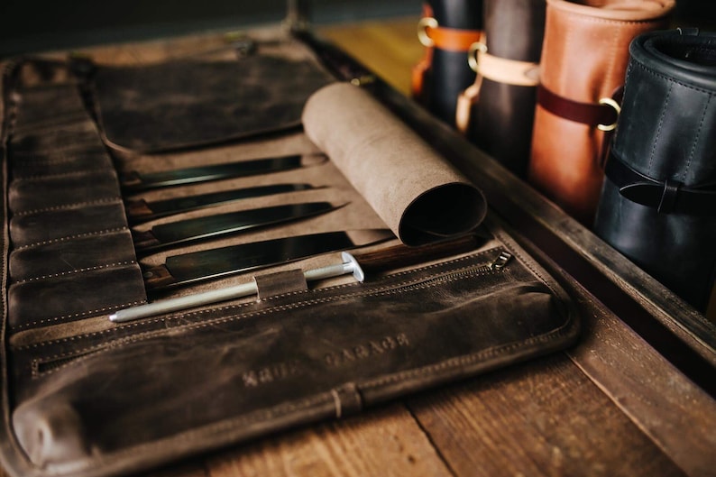 May include: A brown leather knife roll with a zipper closure and a rolled flap. The roll is open and contains several knives and a sharpening steel. The roll is labeled "KRUDGE".